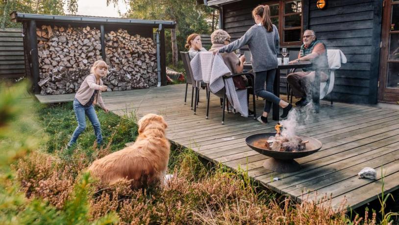 Familie hygger på terrassen i et feriehus i Kystlandet og pige leger med hund og ild i bålfad