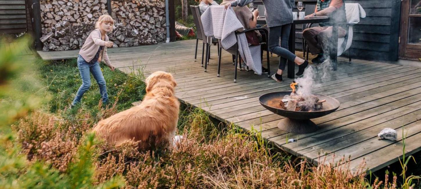 Familie hygger på terrassen i et feriehus i Kystlandet og pige leger med hund og ild i bålfad