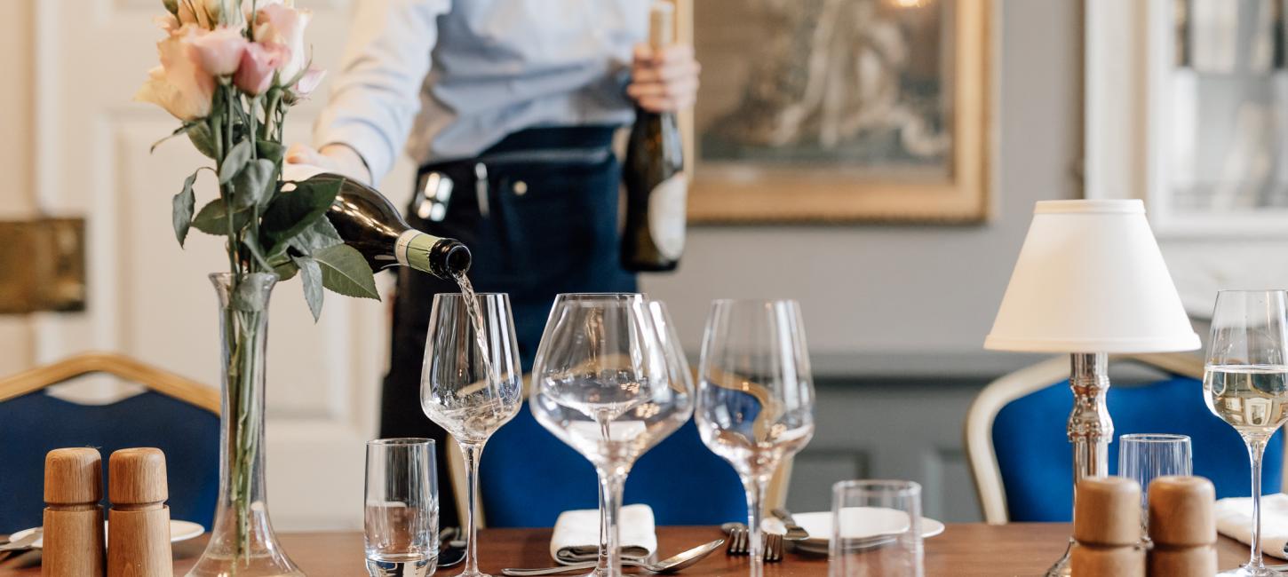 En tjener i blå skjorte hælder vin op i et glas på et flot dækket bord