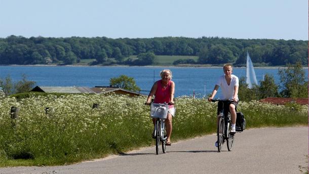 To cyklister cykler langs en vej med vand i baggrunden