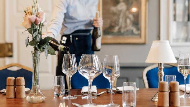 En tjener i blå skjorte hælder vin op i et glas på et flot dækket bord 